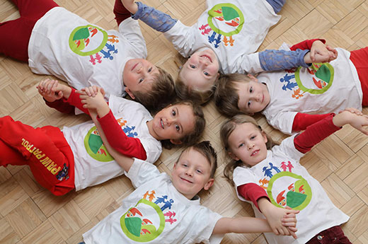 Sechs Kinder mit gleichem T-Shirt liegen auf dem Rücken am Boden und lächeln in die Kamera